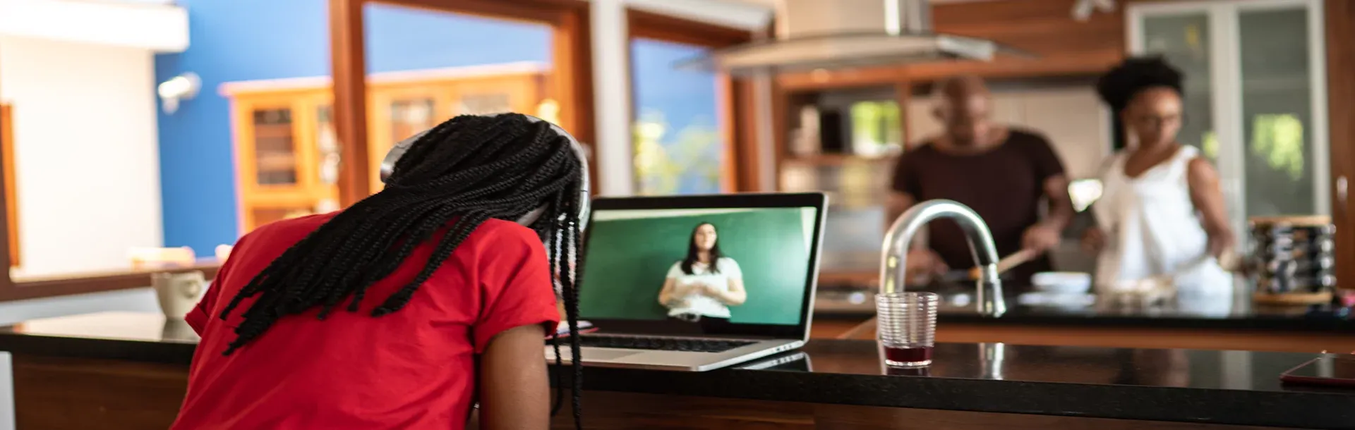 Woman sitting in the kitchen on her laptop taking an online class while someone cooks in the background