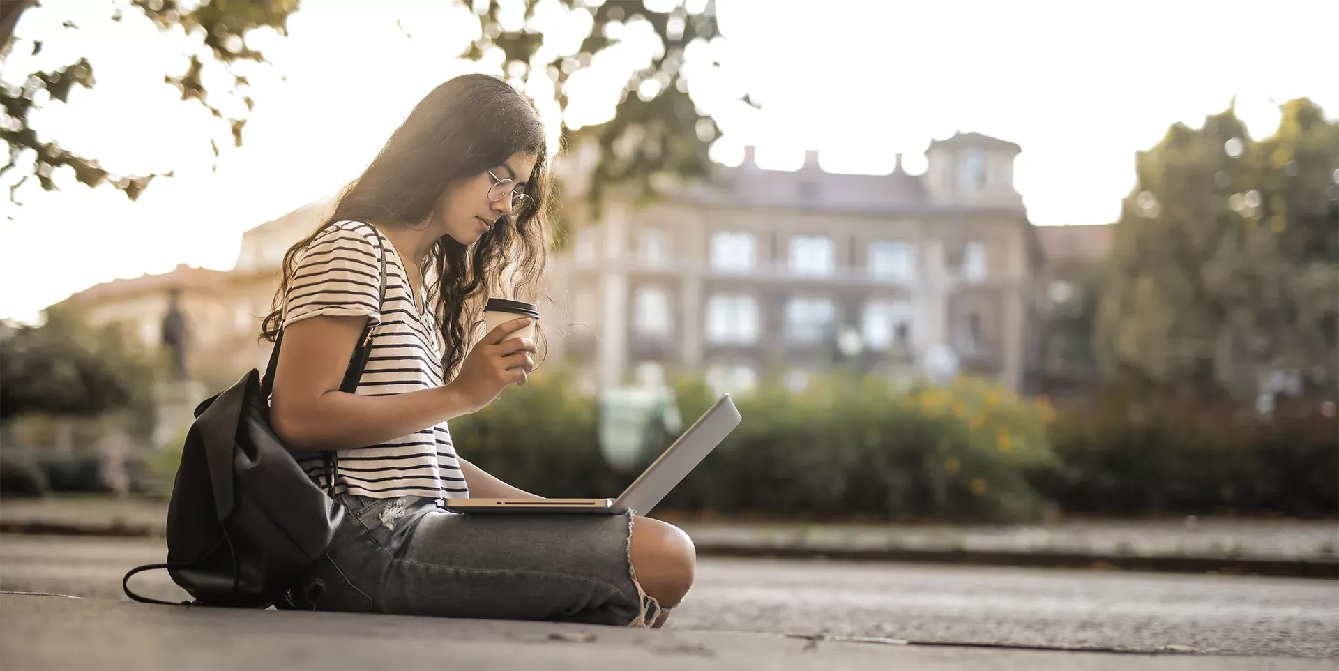 woman sitting on the ground on campus on her laptop while drinking coffee